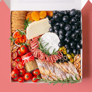 A white square box is packed to the brim with a soft cheese wheel and semi-firm cheese block accompanied by black grapes, cherry tomatoes, green olives, black olives, smoked salmon, turkey rolls, jar of wholegrain mustard, dried apricots, roasted almonds, walnuts, crackers, and baguette slices.