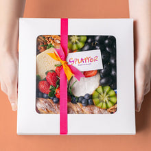 Load image into Gallery viewer, An array of Splatter&#39;s Cheese &amp; Fruit Platter ingredients neatly arranged inside a white square box. The box is covered and contents peek through its transparent window. Two hands hold the platter box sides.