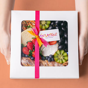 An array of Splatter's Cheese & Fruit Platter ingredients neatly arranged inside a white square box. The box is covered and contents peek through its transparent window. Two hands hold the platter box sides.