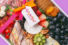 Load image into Gallery viewer, A close-up shot of Splatter&#39;s signature cheese platter covered with a transparent cover tied with pink and orange ribbons, with a white tag branded with Splatter logo. Cheese platter ingredients are visible through the cover.