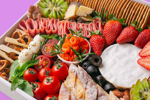 A close-up shot of the cheese platter in a white box, focusing on a round cheese wheel, fresh strawberries, cherry tomatoes, smoked salmon, turkey rolls, caprese salad skewers, and a row of water crackers.