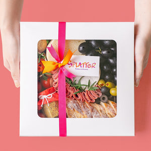 Splatter's Cheese & Meat Platter in a covered white box with transparent window that shows most of its content. Pink and orange ribbons form a knot in the middle with a tag branded with Splatter's logo. Two hands hold the side of the box.