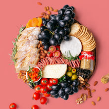 Load image into Gallery viewer, A round wooden board carries a platterful of soft cheese wheel and semi-firm cheese block accompanied by black grapes, cherry tomatoes, green olives, black olives, smoked salmon, turkey rolls, jar of wholegrain mustard, dried apricots, roasted almonds, walnuts, crackers, and baguette slices.