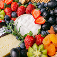 Load image into Gallery viewer, A cheese platter close-up photo featuring a white soft cheese wheel surrounded by fresh strawberries, blueberries, grapes, kiwi, dried apricots, raspberry jam, and a row of water crackers.