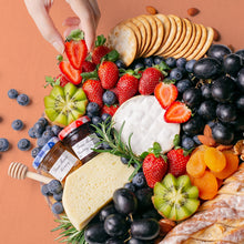 Load image into Gallery viewer, A single hand reaches out to a halved strawberry, one of the many elements arranged on a round wooden board. More fresh strawberries are placed on the platter alongside black grapes, cut kiwi, dried apricots, a round cheese wheel, a block of semi-firm cheese, jars of spreads, and crackers. 