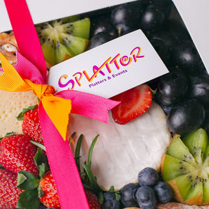 A close-up of Splatter's Cheese & Fruit Platter box with contents peeking through its window cover. Pink and orange ribbons form a knot that carried a branded tag with Splatter's logo.